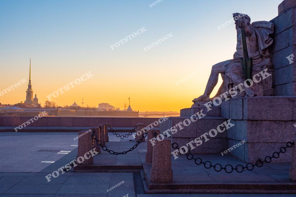 На фотографии запечатлен закатный вид на Петропавловскую крепость с набережной Васильевского острова. На переднем плане — скульптура, украшающая Ростральную колонну, символизирующую морскую славу города. Теплые оттенки заката подчеркивают спокойствие Невы и историческую значимость Петербурга. Этот уголок города является одним из самых узнаваемых и живописных.